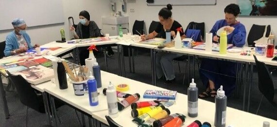 Das Bild ist im Rahmen des onkologischen Kunstprojektes "farbRaum" der Universitätsmedizin entstanden und zeigt diekreative Runde der Teilnehmer*innen. Die Tische sind übersäht von Malutensilien wie Farbtuben und Piinseln.  