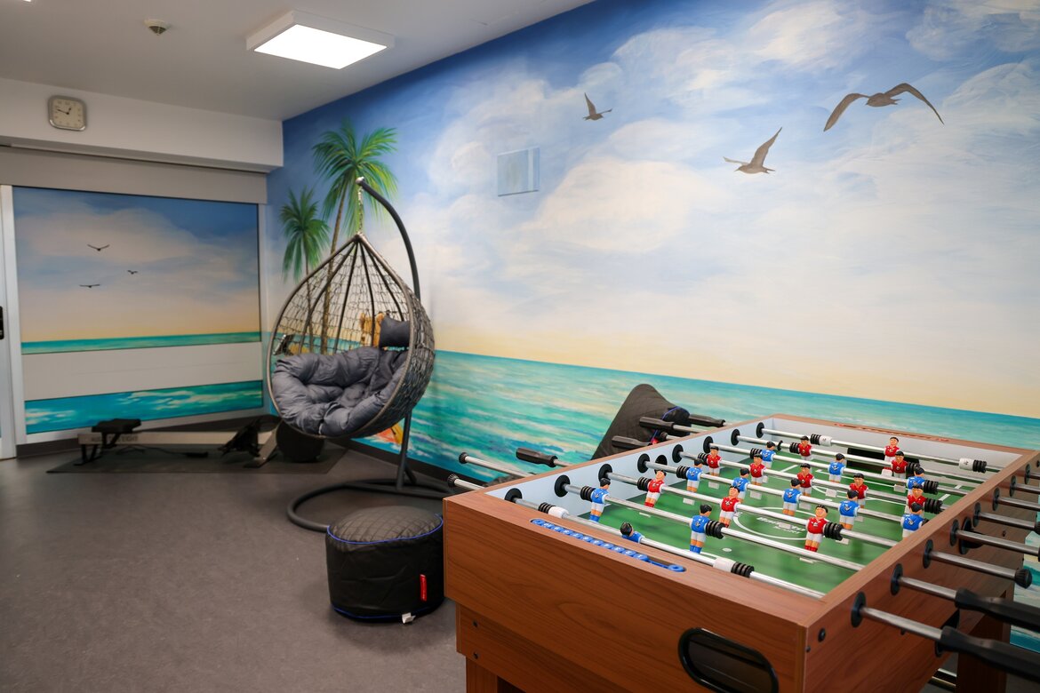 Das Bild zeigt die neu gestaltete Bewegungsinsel der Kinderkardiologischen Station in der Universitätsmedizin Göttingen. Zu sehen ist die liebevoll gestaltete Wandmalerei, die eher an einen Aufenthalt am Meer als im Krankenhaus erinnert. Davor stehen ein gemütlicher Korbsessel, der Kicker und ein Rudergerät. 