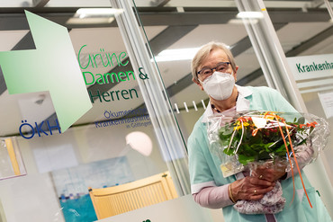 Irene Tuschhoff „Grüne Dame“ der ersten Stunde, arbeitet seit 40 Jahren ehrenamtlich an der UMG. Sie steht vor dem Büro mit der Aufschrift "Grüne Damen und Herren" mit dem typischen grünen Kittel, Mundschutz und einem Blumenstrauß. Foto: Foto: UMG/spförtner 